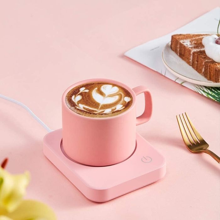 A pastel pink mug warmer sitting on a desk with a coffee mug resting on top
