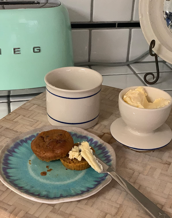 A white butter cup with a lid, decorated with blue lines, placed next to a buttered muffin.