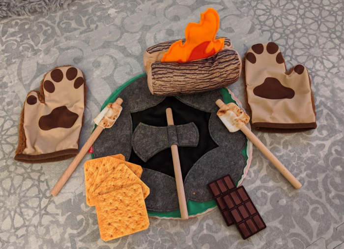 A reviewer's photo showing a felt campfire playset, complete with a pair of bear hands, a burning log, an axe, and s'mores, ready for imaginary adventures.