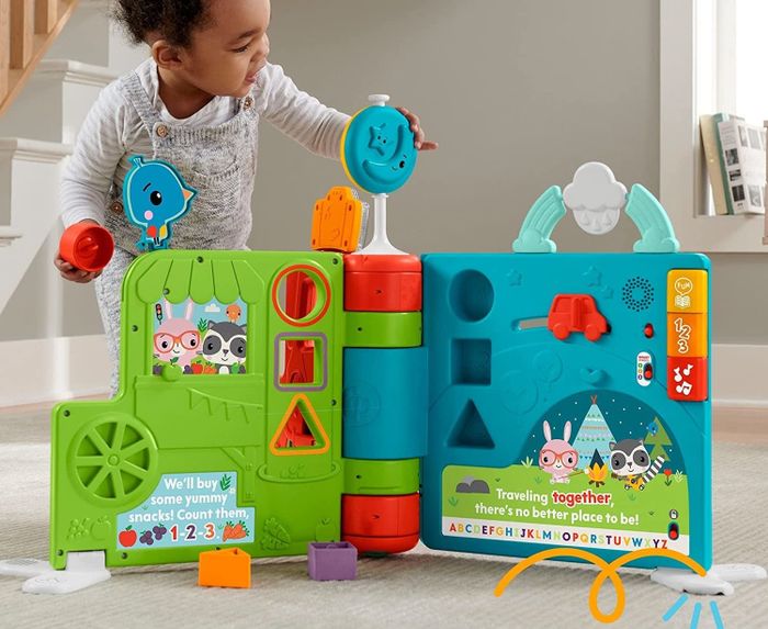 A young child standing behind a colorful plastic toy book, with the pages open to show the activities inside