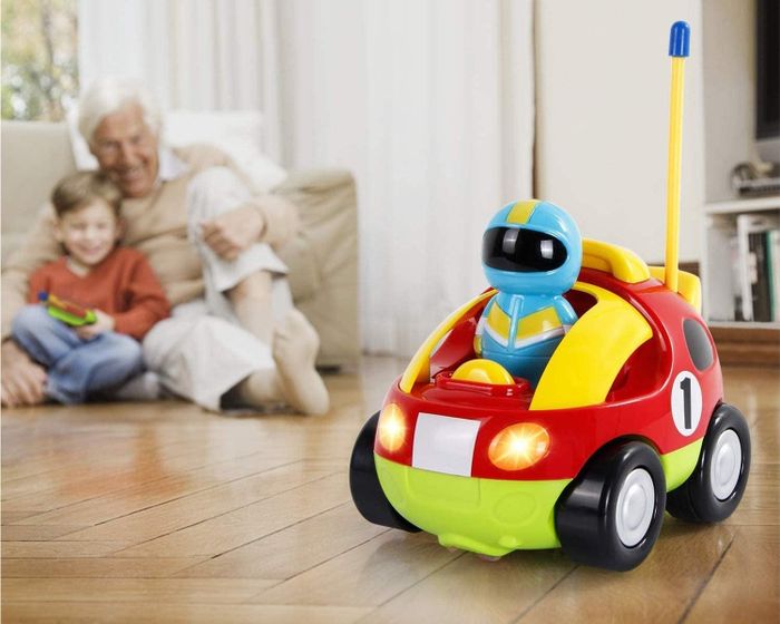 A vivid red, green, and yellow plastic race car toy, being operated by a child model while an adult model supervises the action