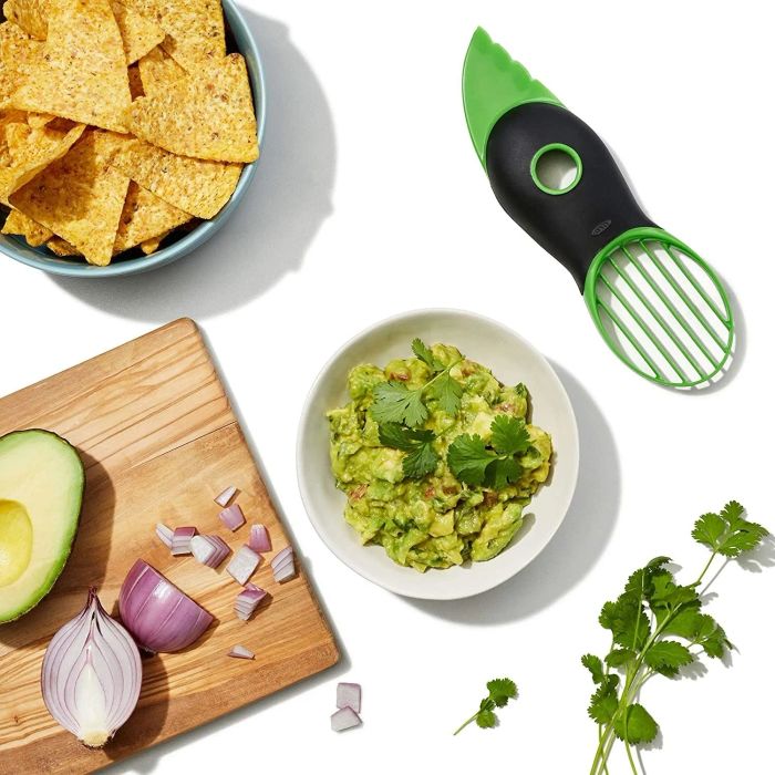 A green avocado slicer placed next to a bowl of guacamole and slices of avocado on a cutting board