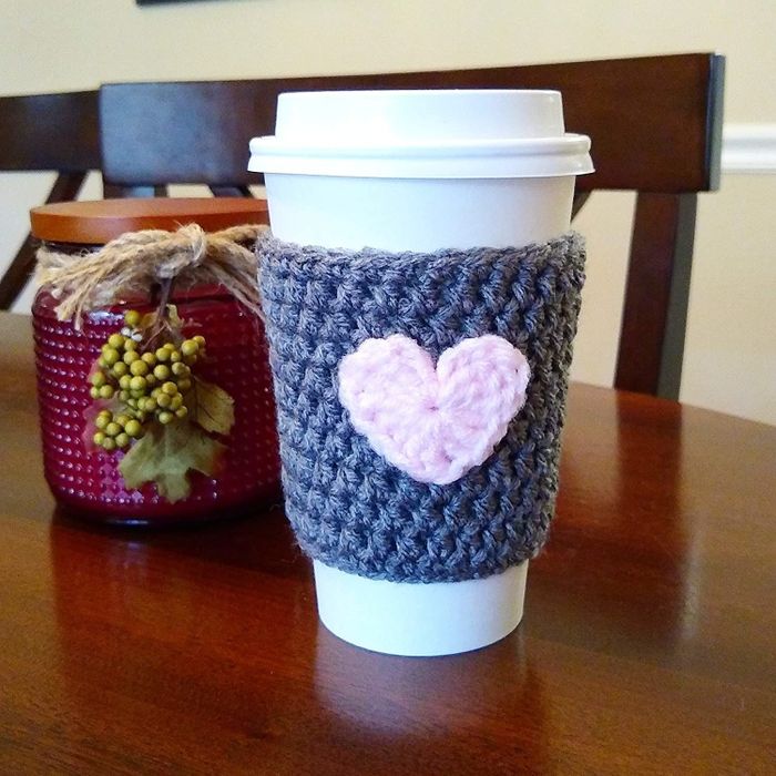 A white disposable coffee cup with a gray knitted sleeve featuring a soft pink heart in the center.