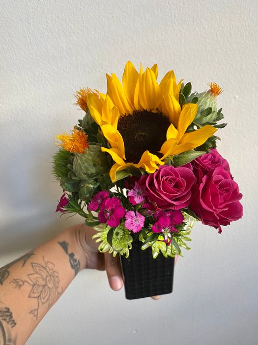 A model’s hand holding a bouquet with pink roses, sunflowers, and other flowers.