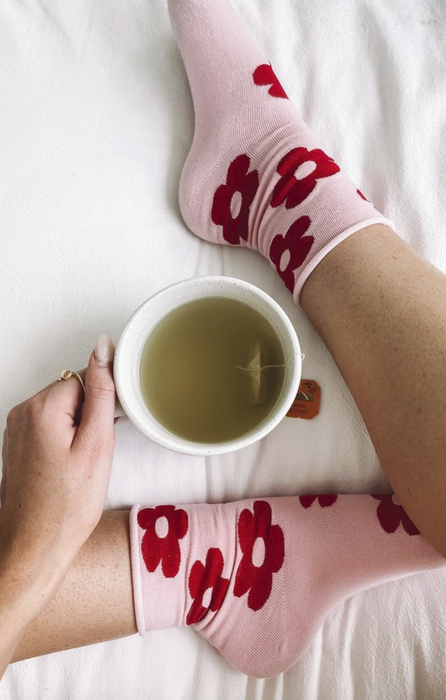 Close-up of a model's feet wearing light pink socks adorned with red flowers, with a cup of coffee placed between them.