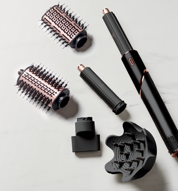 A variety of hair styling tools, including a blow dryer, curling wands, and brushes, arranged on a marble surface.