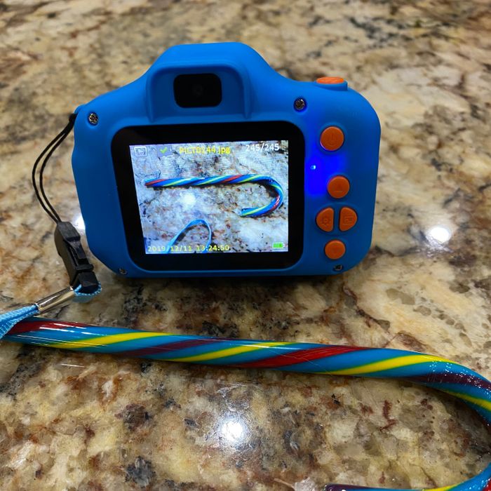 A toy camera shows a rainbow candy cane on its display; the candy cane matches one placed beside it on a stone countertop.