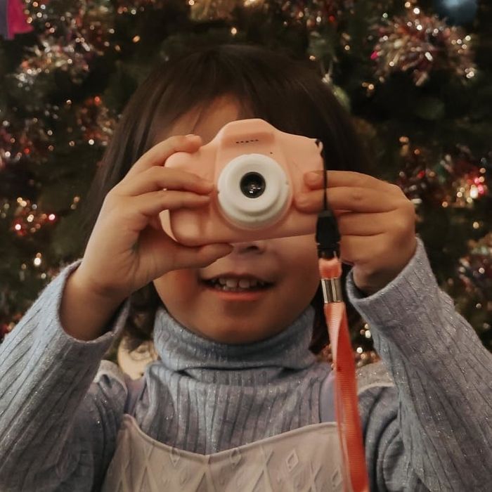 A child stands in front of a Christmas tree, holding a pink camera, dressed in an Elsa-themed dress from Frozen.