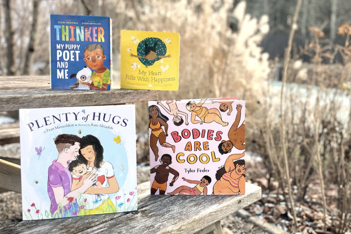 Displayed on a wooden surface, a selection of children's books includes 'Thinker: My Puppy Poet and Me' by Eloise Greenfield, 'My Heart Fills with Happiness' by Monique Gray Smith, 'Plenty of Hugs' by Fran Manushkin, and 'Bodies Are Cool' by Tyler Feder.