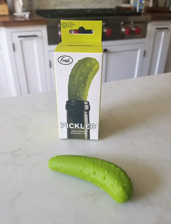 A pickle-shaped wine stopper, placed next to its packaging, adding a fun and unique twist to your wine accessories.