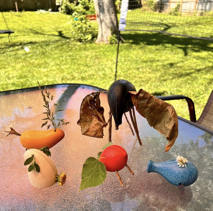 Intricate fruit and vegetable sculptures incorporating natural elements like leaves and flowers, carefully arranged on a glass surface outdoors.