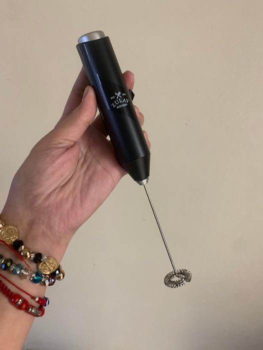 A hand gripping an electric milk frother, its wrist decorated with colorful bracelets.