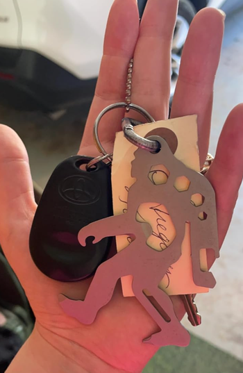 A hand grips a set of keys, adorned with a Bigfoot bottle opener keychain, a Toyota fob, and a labeled key.
