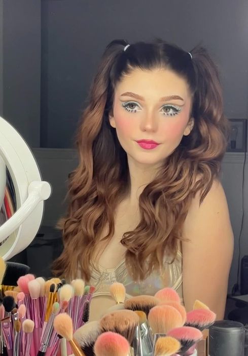 A reviewer rocking colorful winged eyeliner and two playful hair buns, surrounded by her collection of makeup brushes.