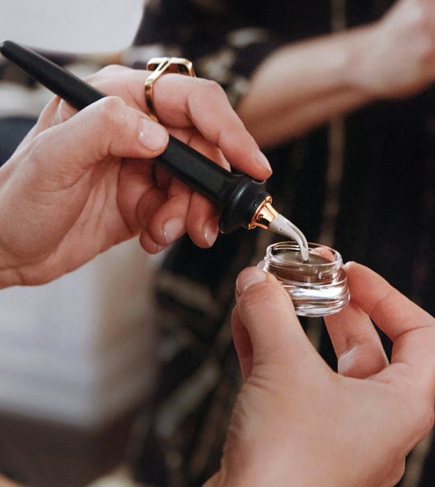 A model dipping the guide wand into a small container of brown eyeliner to prepare for application.