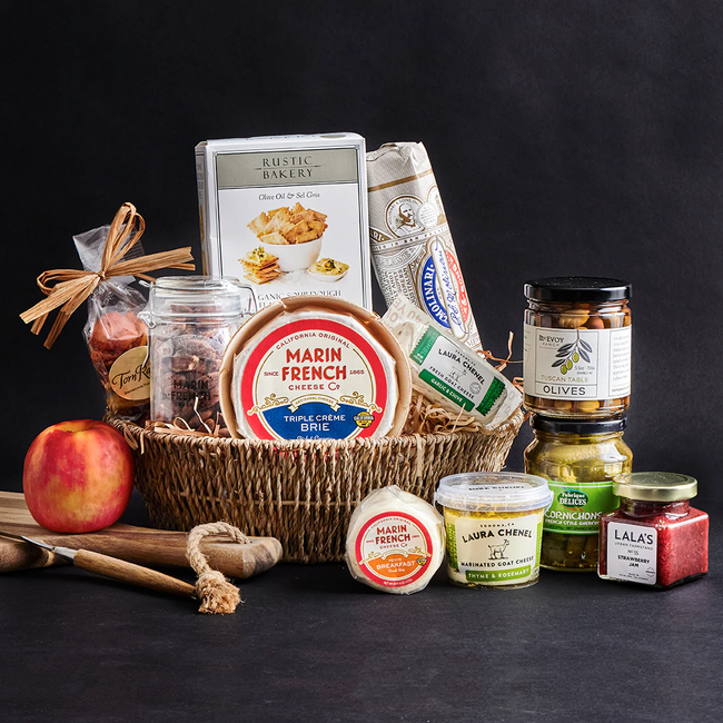 A basket filled with gourmet goodies like cheese, jam, olives, crackers, and tea, along with wooden serving utensils and a fresh apple