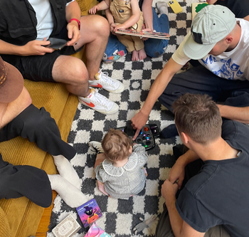 A group of people gathered around a baby playing on a checkered rug, surrounded by toys and books.