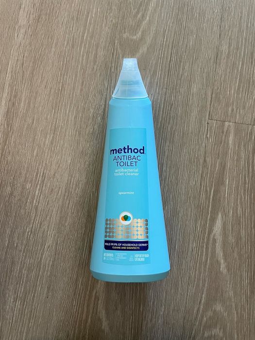 A bottle of Method antibacterial toilet cleaner on a wooden floor, with spearmint scent, emphasizing its germ-killing action.