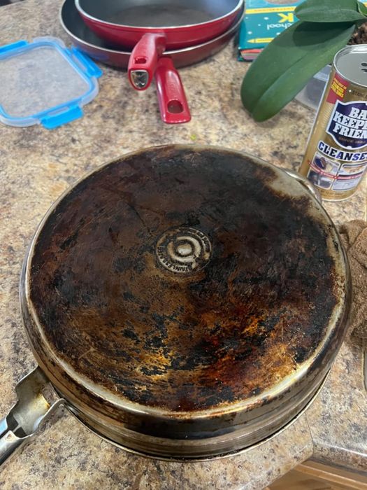 An old pan with a heavily burnt base sits on a kitchen counter, surrounded by cleaning supplies and a red pan.