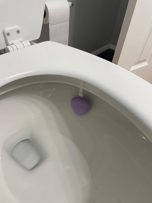 A toilet bowl featuring a lavender-scented cleaner attached inside, with a small visible portion of a toilet paper roll nearby.