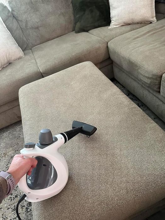 A hand uses a steam cleaner on a fabric-covered ottoman in a cozy living room.