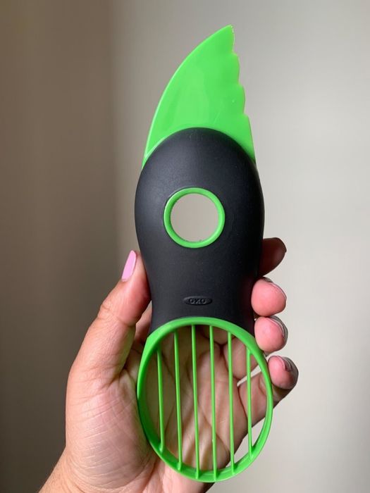 a handle gripping the green and black avocado slicer, featuring a serrated blade at the top, a round pit remover in the center, and a fan blade to slice the avocado into seven pieces