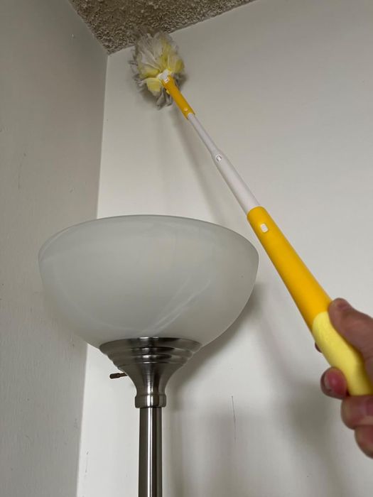 A person using a yellow extendable duster to clean the corner of a ceiling next to a standing lamp.