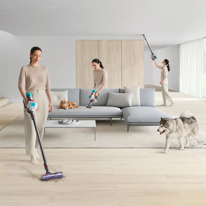 A woman vacuums a modern living room while her dog watches nearby. Dressed in stylish neutral attire, she demonstrates efficient home cleaning.