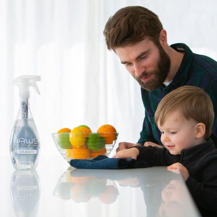An adult and child clean a table with a spray bottle of Yaws Granite cleaner placed nearby, alongside a bowl filled with oranges and limes