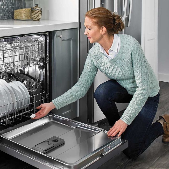 A person wearing a sweater loads plates into a dishwasher and adds a detergent tablet, showcasing the appliance’s functionality in a home kitchen setting
