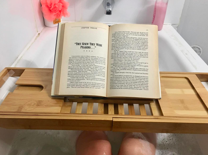a reviewer’s photo of someone in a bathtub using the tub caddy to hold their book, keeping it dry while they read during their bath