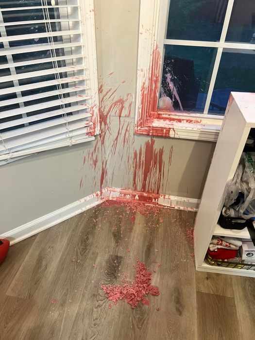 Pink wax has been spilled and splattered across the corner of a room, covering the floor and window ledge. Some of the wax has gathered in a pool on the floor.