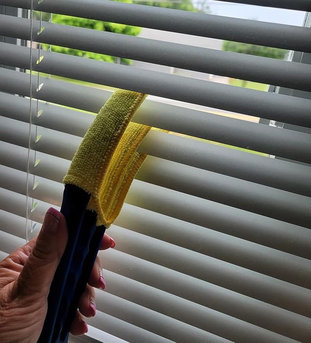 A person is seen using a cleaning tool to dust a set of horizontal blinds.