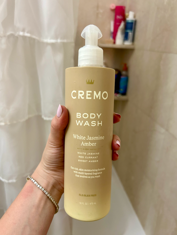 A hand holding the Cremo Body Wash bottle, White Jasmine Amber scent, in a bathroom with a shower curtain and toiletries visible in the background