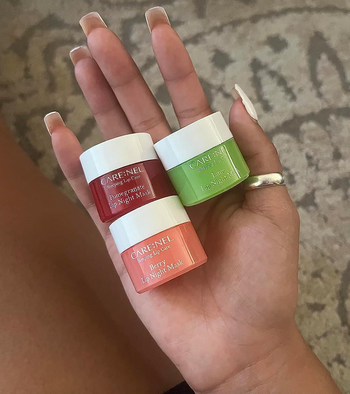 A reviewer holds three small containers of lip mask in berry, pomegranate, and lime flavors.