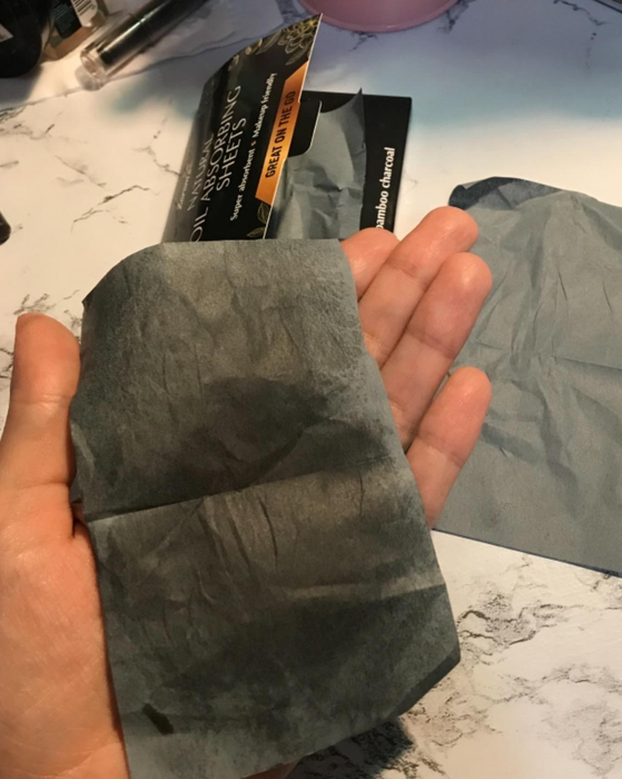 A hand holding opened sheets of charcoal blotting papers, with a used sheet and the packaging displayed nearby on a marble surface.
