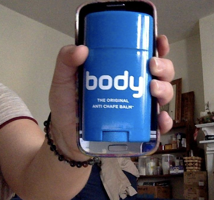 A reviewer holding a stick of anti-chafe balm in front of an iPhone, demonstrating that the product is roughly the same size as the phone.