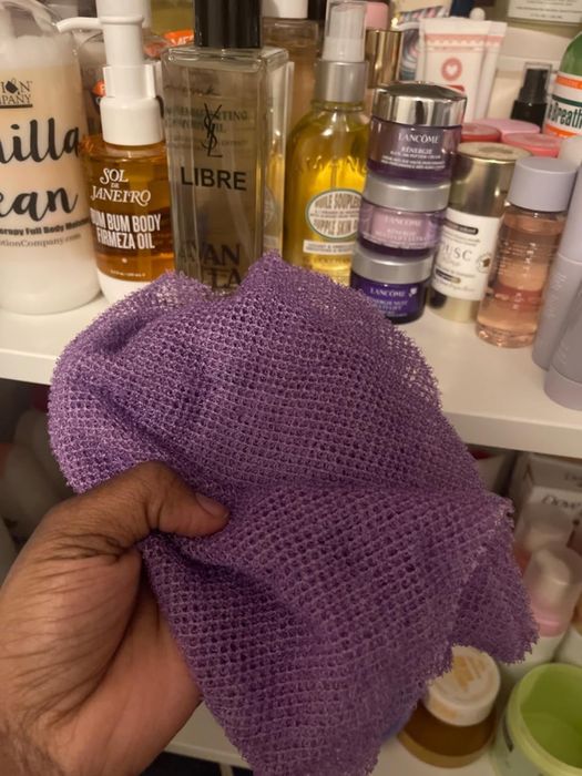 A hand holding a purple exfoliating cloth in front of a shelf filled with various skincare and beauty products in bottles and jars.