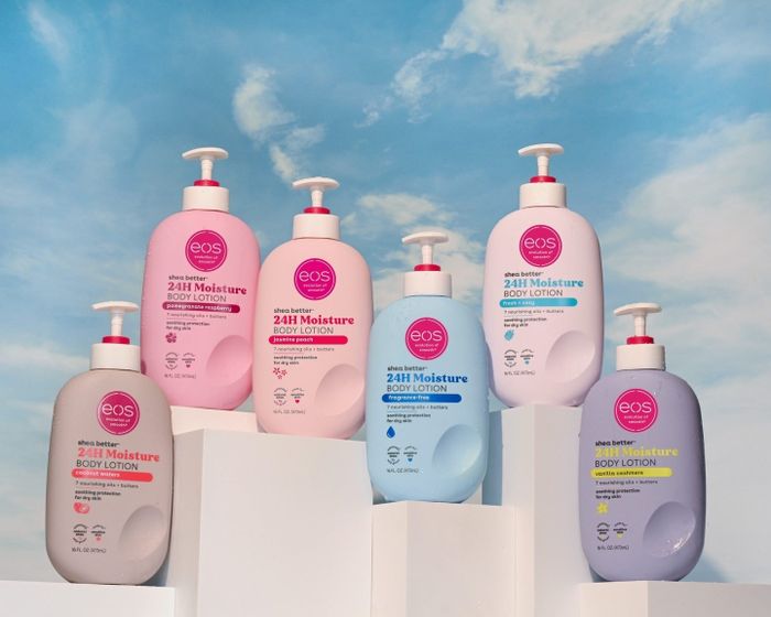 A display featuring six EOS 24H Moisture Body Lotion bottles in a variety of scents, arranged on white pedestals with a sky backdrop.