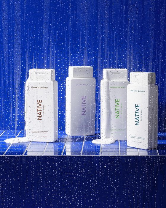 Four bottles of Native body wash resting on a tiled surface, with droplets of water, featuring scents such as coconut & vanilla, lavender & rose, and sea salt & cedar.
