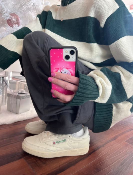 A person sitting cross-legged, holding a phone with a pink case, wearing a green and white striped sweater and beige sneakers