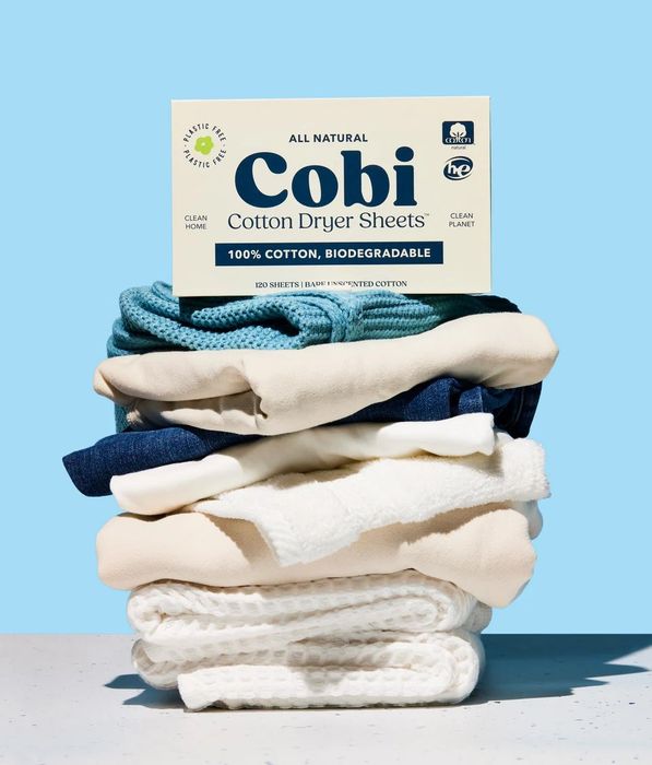 A stack of folded natural fiber fabrics with a box of Cobi cotton dryer sheets on top, labeled biodegradable and 100% cotton