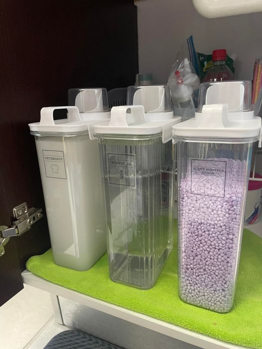 Three labeled containers for storing laundry detergent, softener, and softener powder sheets, placed on a green mat inside a cabinet.