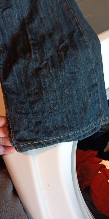 A hand holding a wrinkled, possibly soiled pair of jeans over the door of a washing machine full of clothes.