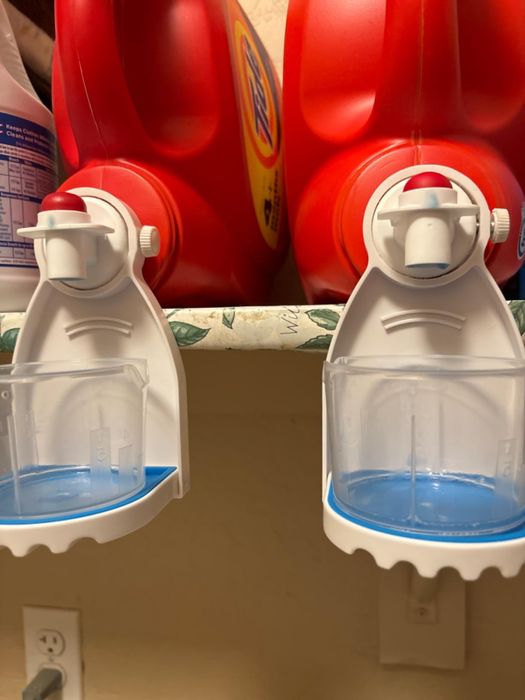 Laundry detergent dispensers attached to large detergent containers on a shelf.