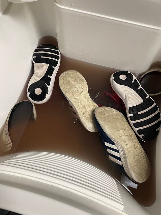 Four shoes and a pair of sunglasses submerged in muddy water inside a soaking tub.