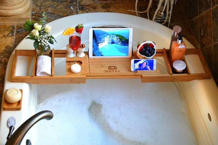 A wooden tray resting over a bathtub, complete with an iPad, a glass of wine, fresh flowers, and a collection of bath scrubs.