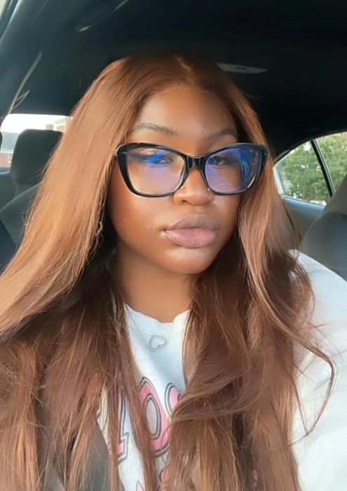 A person with long hair and large glasses is sitting in a car, wearing a casual outfit and gazing directly at the camera.
