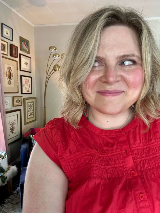 A person smiling in an art-filled room, dressed in a ruffled, sleeveless top.