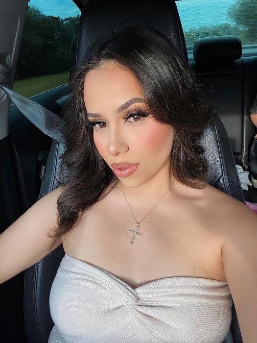A woman sitting in a car, wearing a strapless top and a cross necklace, with long, wavy hair, gazing at the camera with a neutral expression.